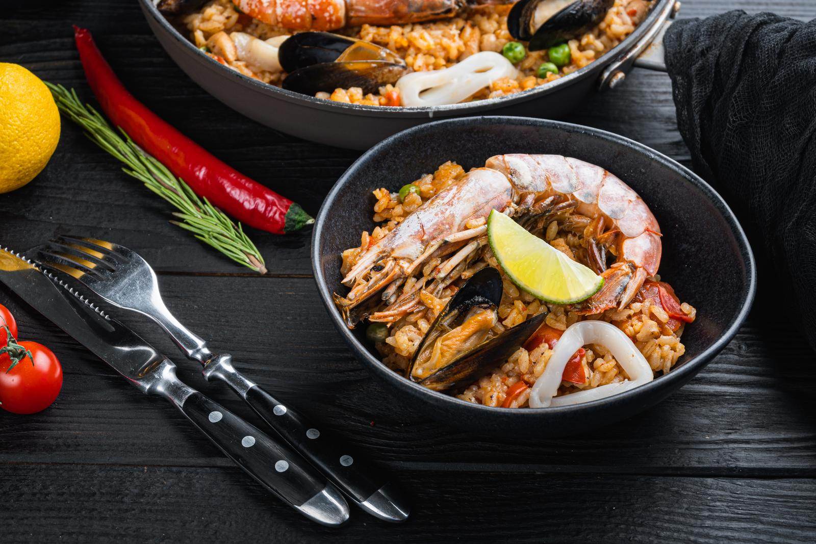 Traditional Spanish Seafood Paella In Pan And Bowl With Rice, Peas, Shrimps, Mussels And Squid On Black Wooden Planks